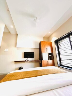 Habitación de hotel con cama, televisor montado en la pared, aparador de madera y ventana con persianas.