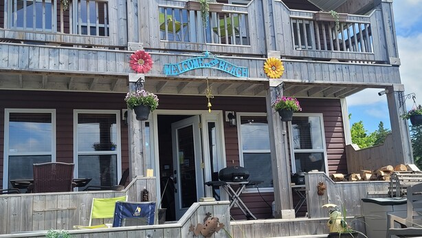 Edificio de madera de dos pisos con un balcón, un cartel que dice 'Welcome to the Beach' y mobiliario exterior con una parrilla.