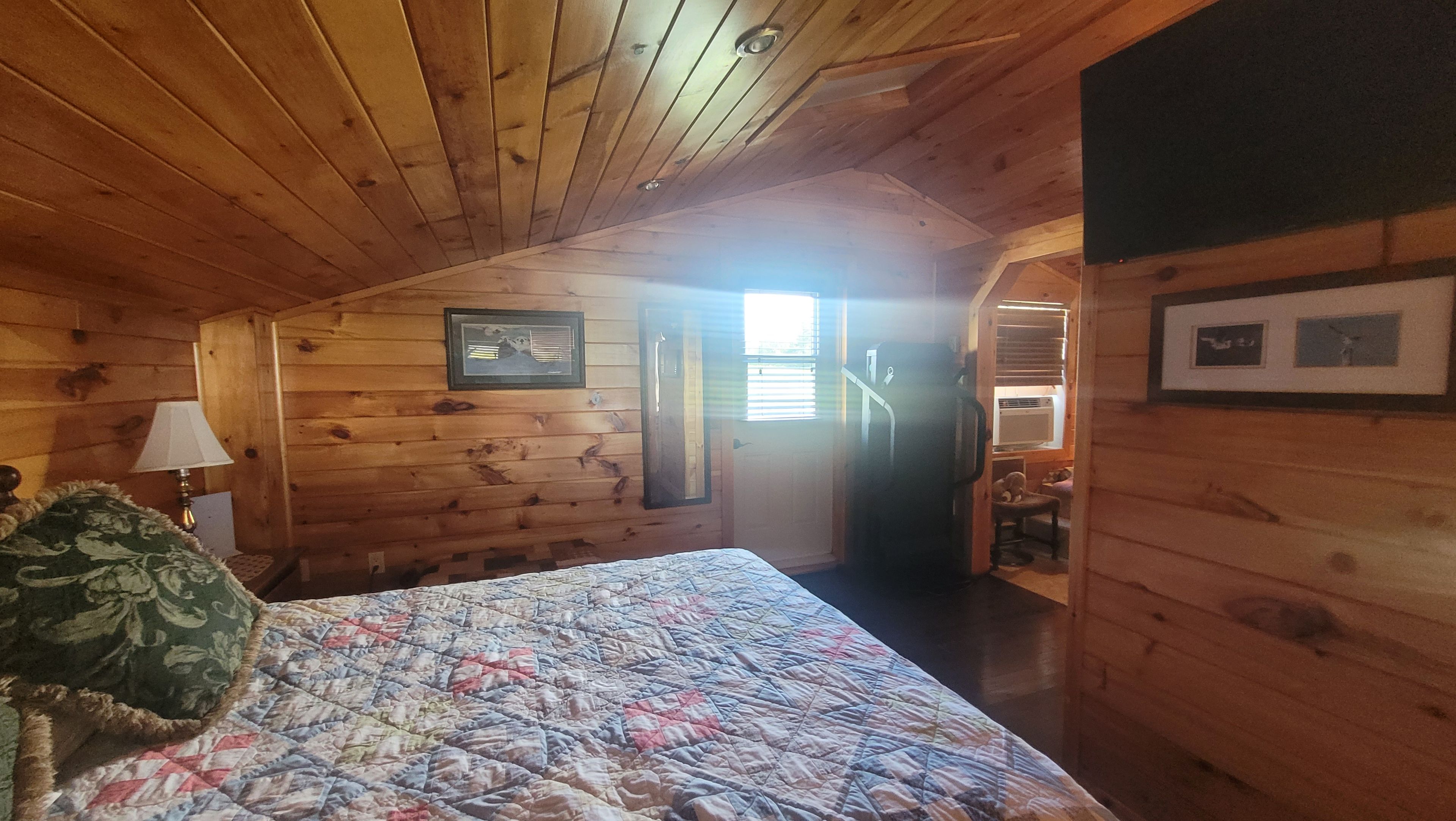 A wooden cabin bedroom with a bed, a nightstand, a lamp, and a door leading to a bathroom.