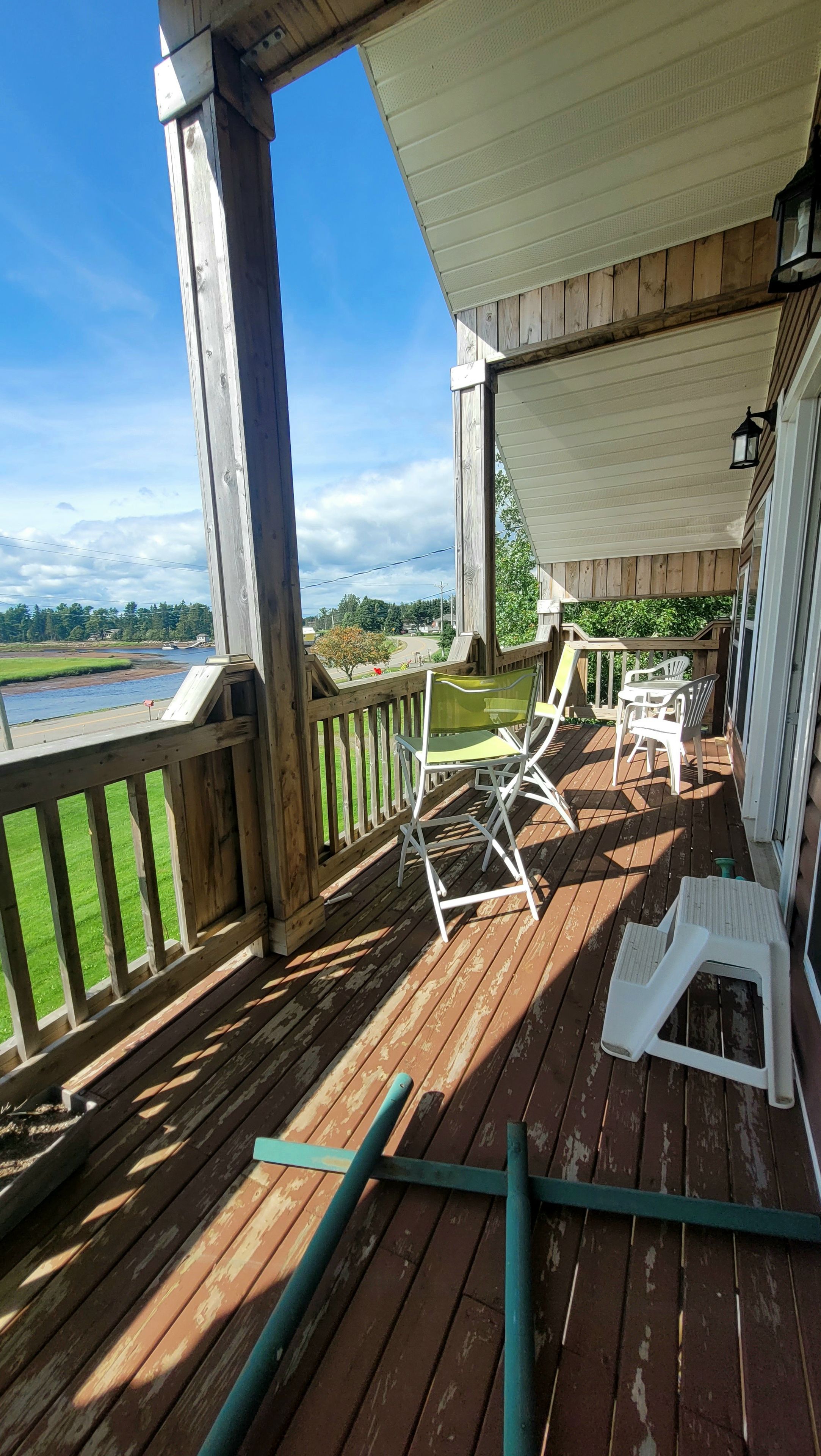 Une terrasse en bois offrant une vue sur une rivière et la verdure environnante, des chaises en plastique blanches et un espace couvert avec une table.