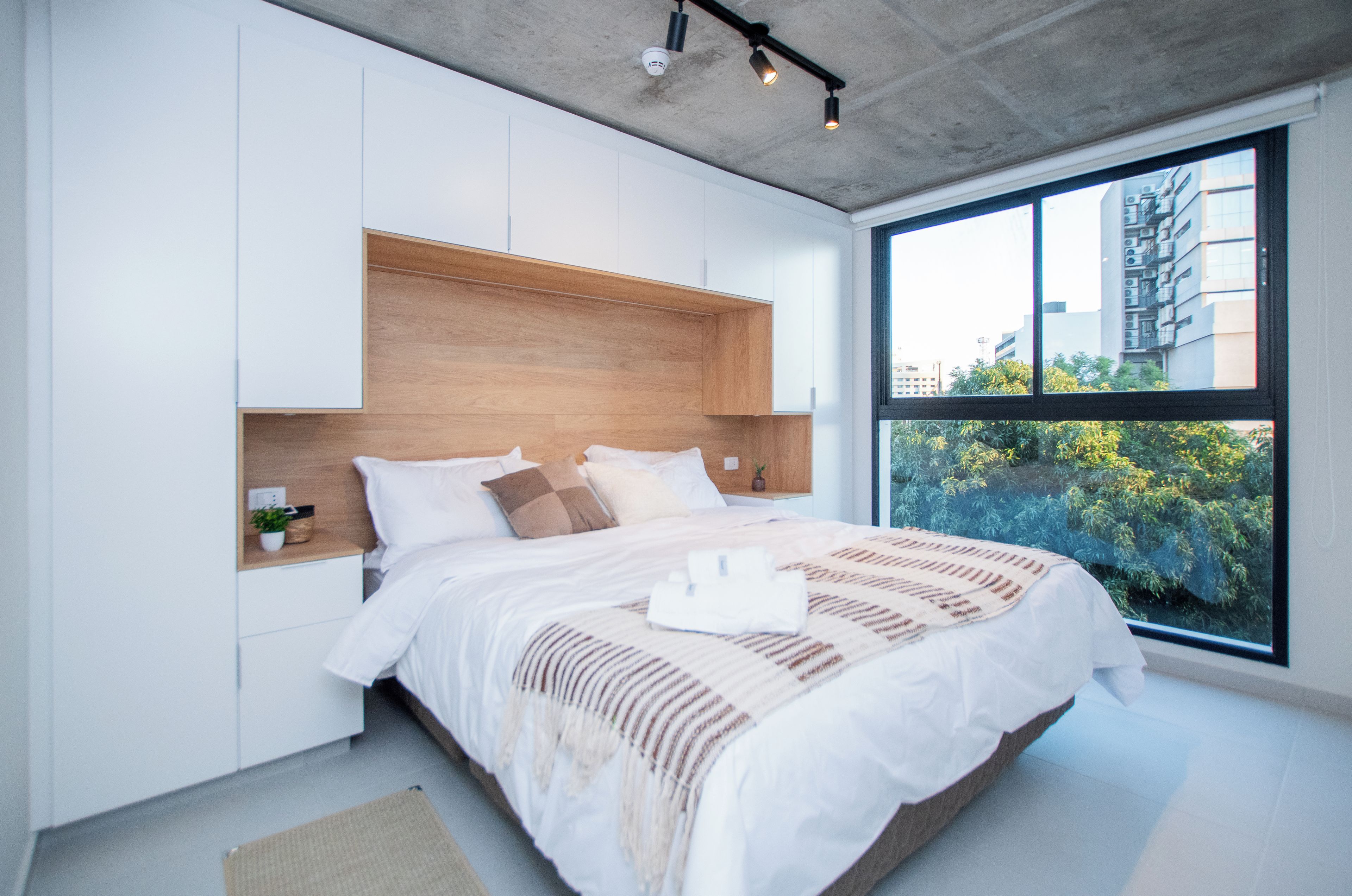 Une chambre moderne avec un grand lit, une tête de lit en bois intégrée, des murs blancs et une fenêtre donnant sur des immeubles et des arbres.