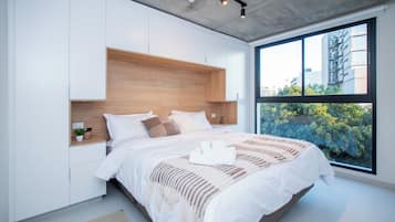 Une chambre moderne avec un grand lit, une tête de lit en bois intégrée, des murs blancs et une fenêtre donnant sur des immeubles et des arbres.