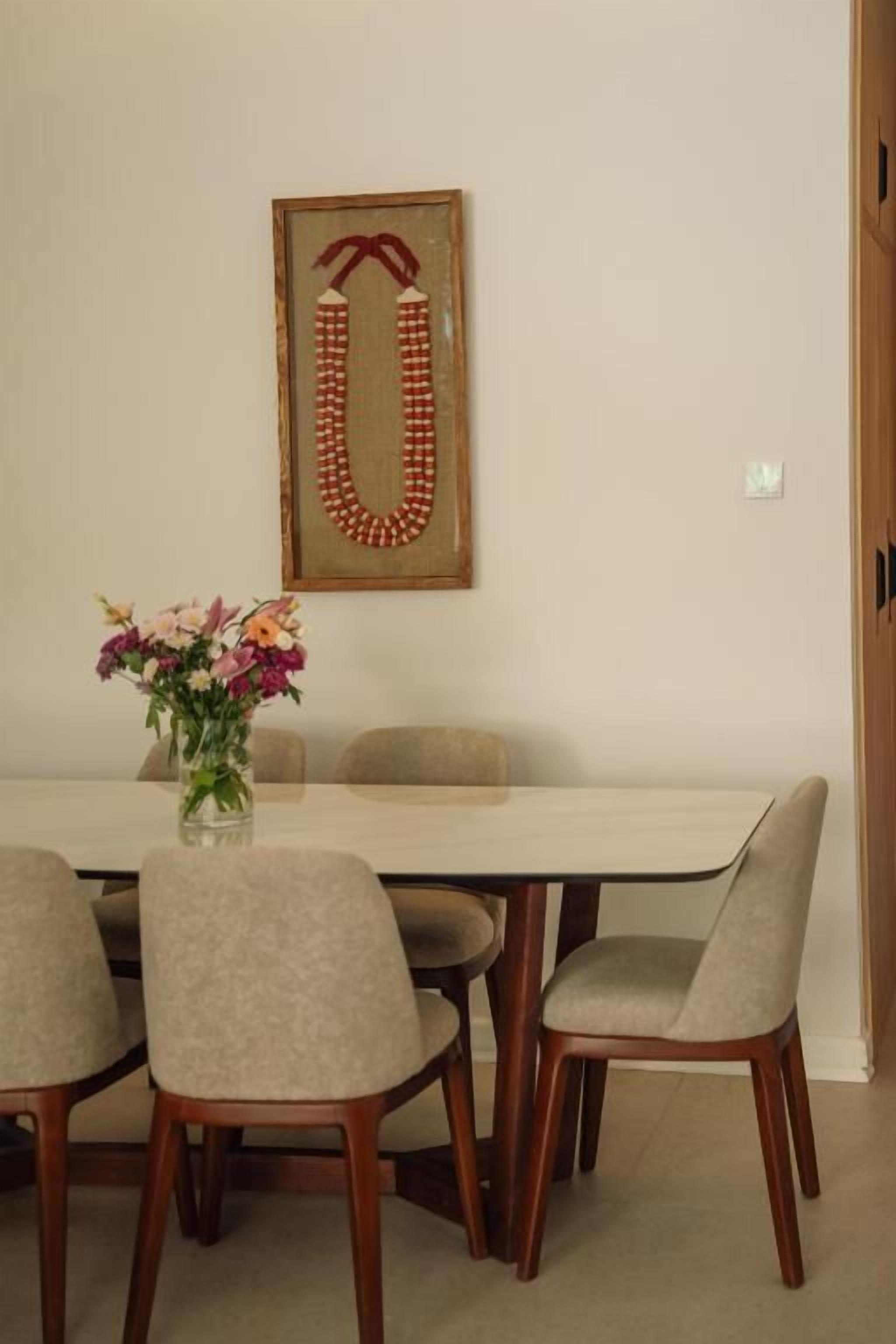 A dining area with a table, chairs, a vase with flowers, and a framed necklace on the wall.
