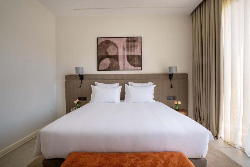Une chambre d’hôtel avec un grand lit, deux tables de chevet, une tête de lit, une œuvre d’art accrochée au mur et une fenêtre avec des rideaux.