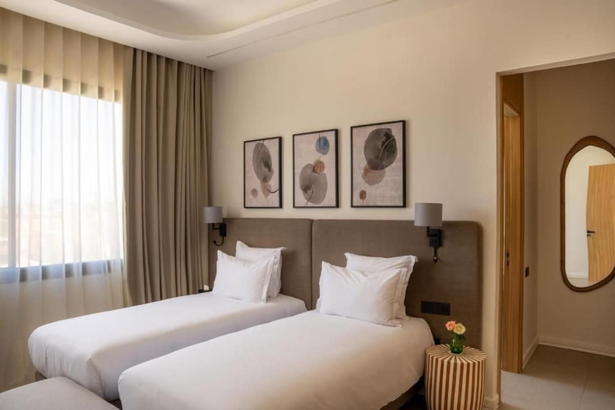 A hotel room with two beds, a large window with curtains, wall-mounted artwork, and a mirror.