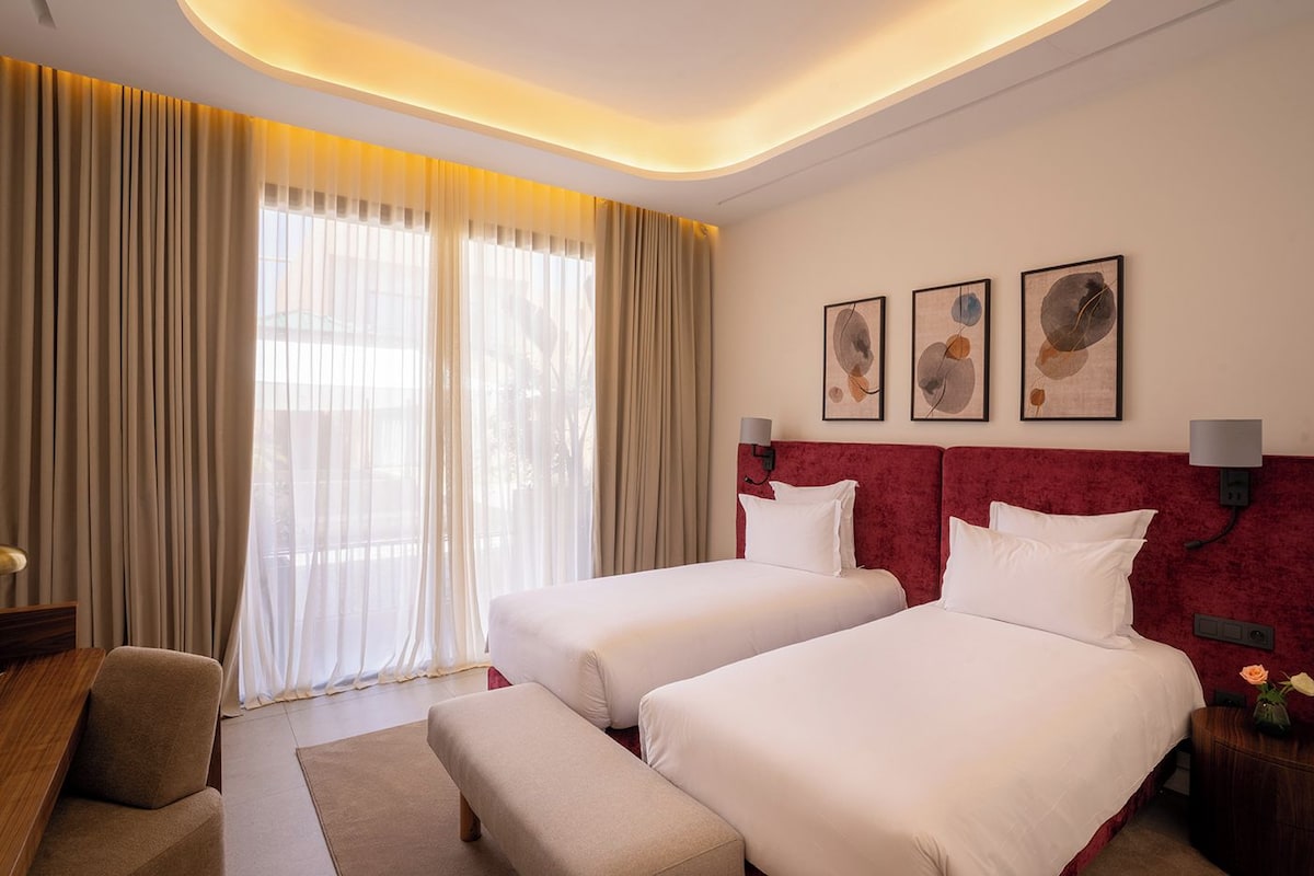 A hotel room with two beds, a desk, a chair, and a large window with curtains.
