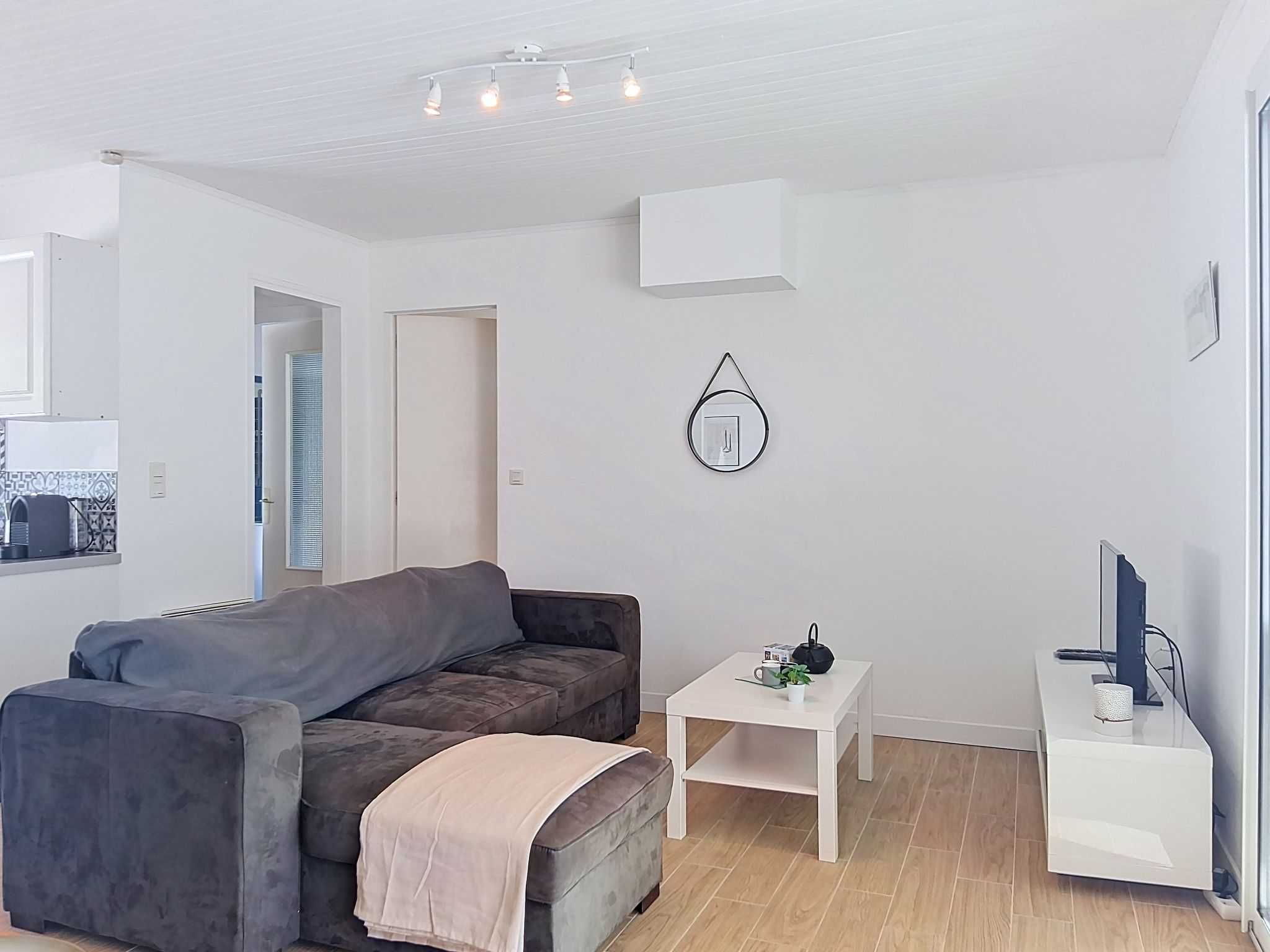 Een moderne woonkamer met een grijze bank, een klein wit bijzettafeltje, een witte kast en een wandlamp.
