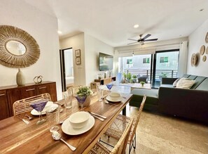 Espaço de refeições com mesa de madeira posta para quatro pessoas, sofá e varanda com plantas em vasos.