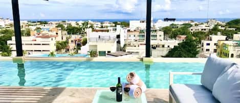 Zona de piscina con vista a un paisaje urbano, una mesa con una botella y vasos, y un sofá.