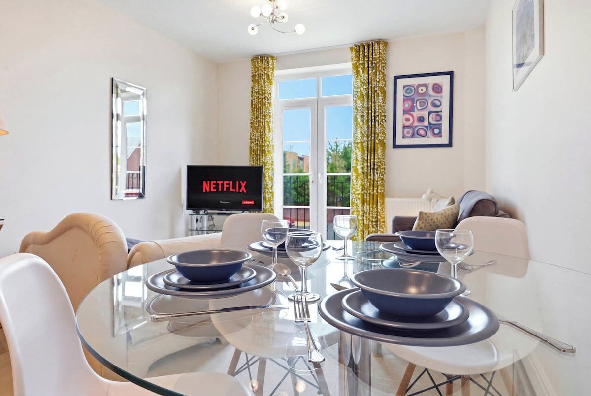 A modern living room with a glass dining table set for four, a sofa, a TV displaying 'NETFLIX', and a window with curtains.