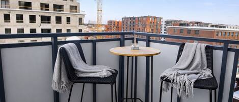 A balcony with two chairs and a small table, overlooking a cityscape with buildings and a crane.