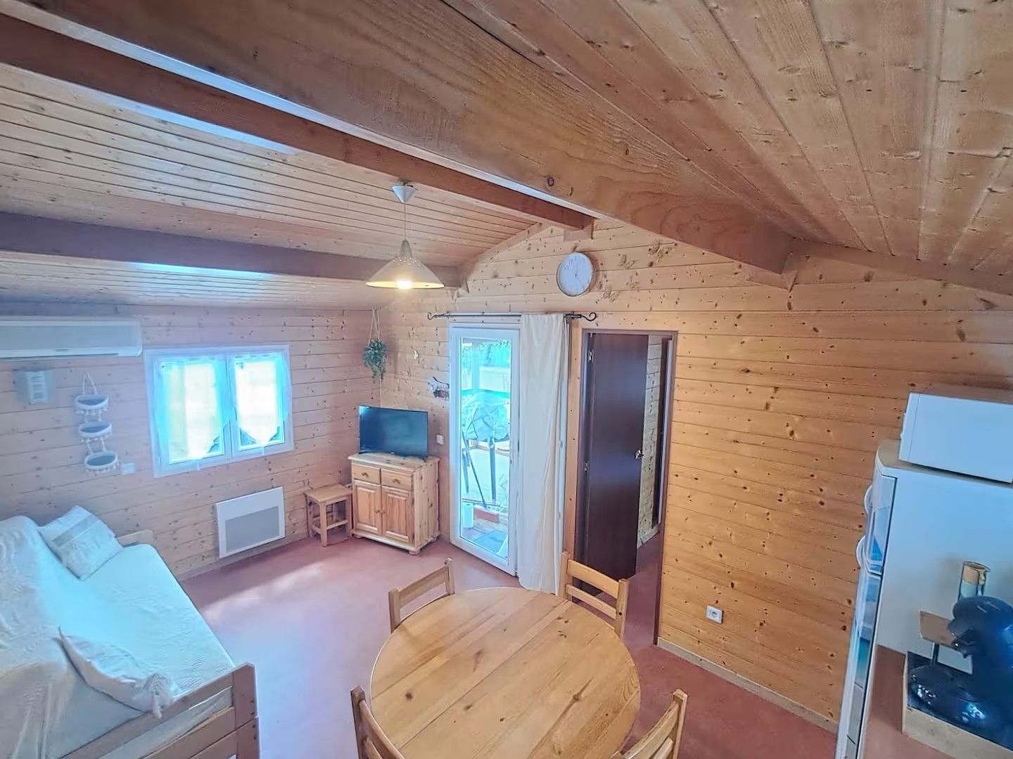 Een houten blokhut interieur met een bed, een eettafel, een televisie en een klok aan de muur.