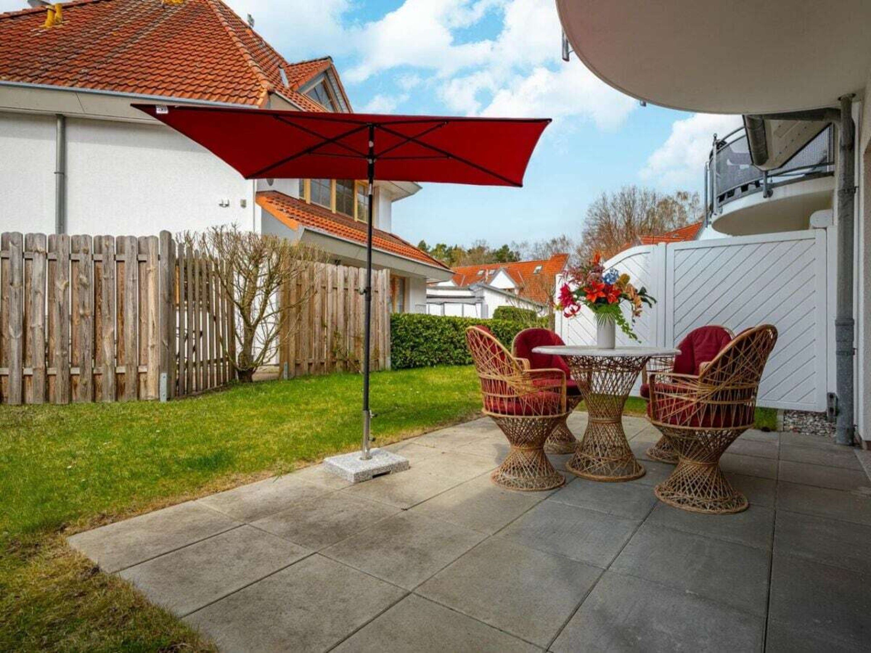 A patio with wicker furniture, a red umbrella, and a white fence.
