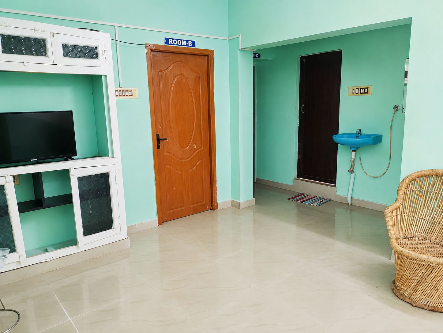 A room with a television on a white cabinet, a wooden door labeled 'ROOM-B', and a blue sink.