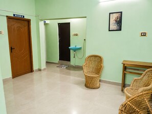 Un pasillo con una puerta de madera que dice 'ROOM-B', un lavabo azul, sillas de mimbre y un cuadro en la pared.