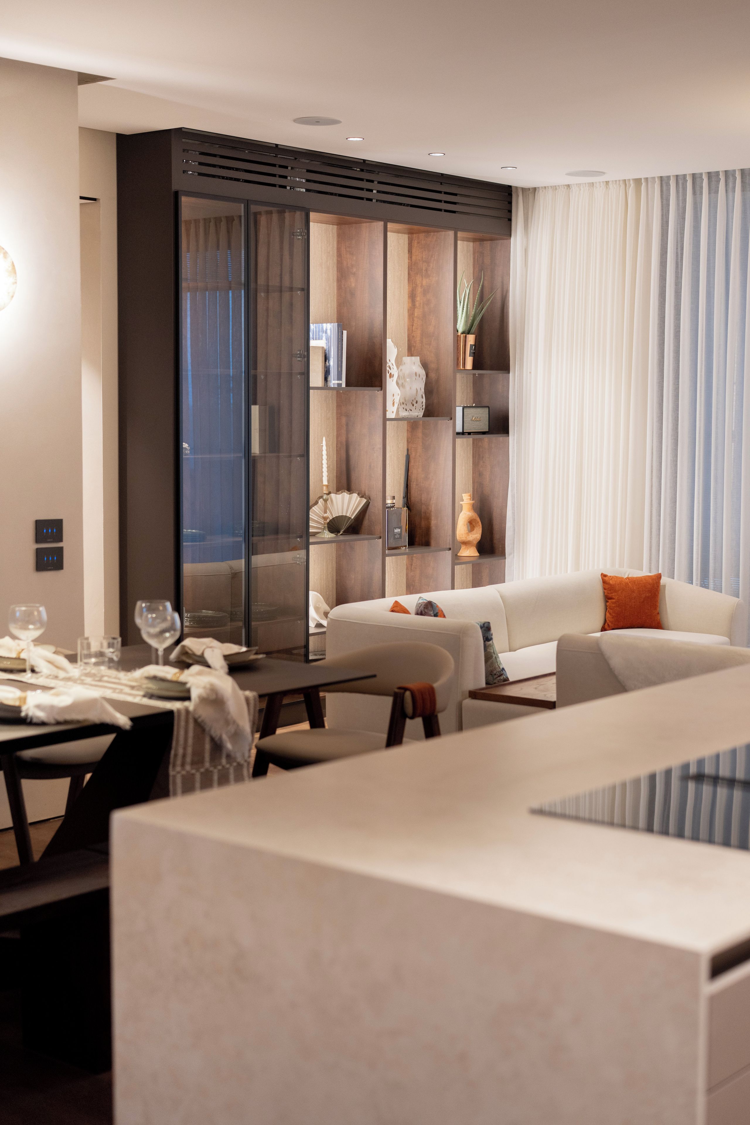 A modern living room with a dining area, featuring a sofa, a dining table set for two, and a glass partition with shelves.