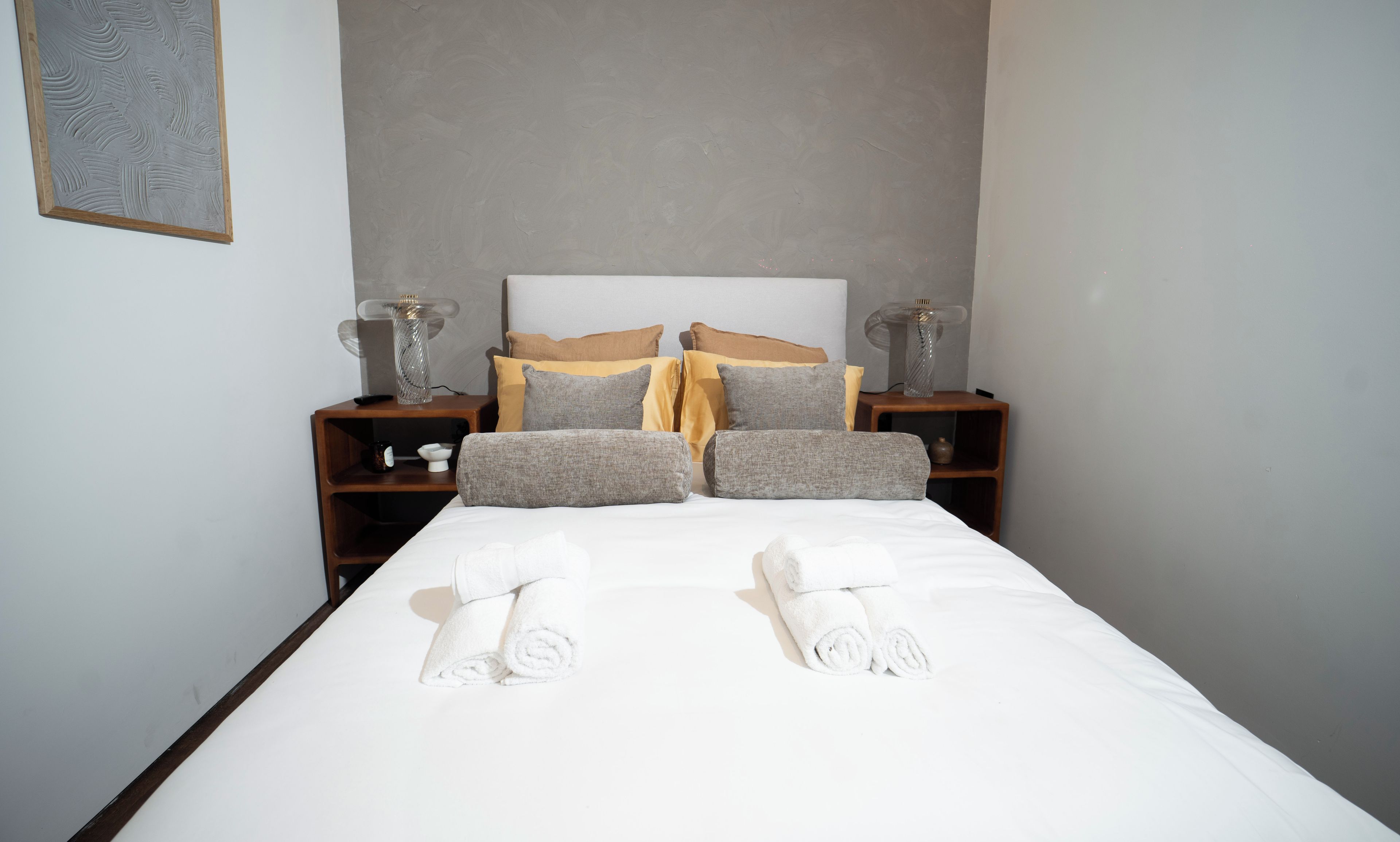 A neatly made bed with white linens and rolled towels, flanked by two nightstands with lamps and a small shelf, against a plain wall.
