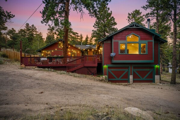 Une cabane à deux étages, dotée d’une véranda et d’un garage, entourée d’arbres.
