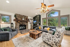 A spacious living room with a stone fireplace, a ceiling fan, and large windows offering a view of the outdoors.