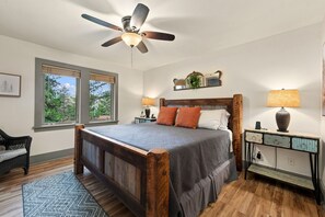 Une chambre à coucher avec un lit en bois, un ventilateur de plafond, une table de chevet, une lampe, une commode et une fenêtre donnant sur des arbres.