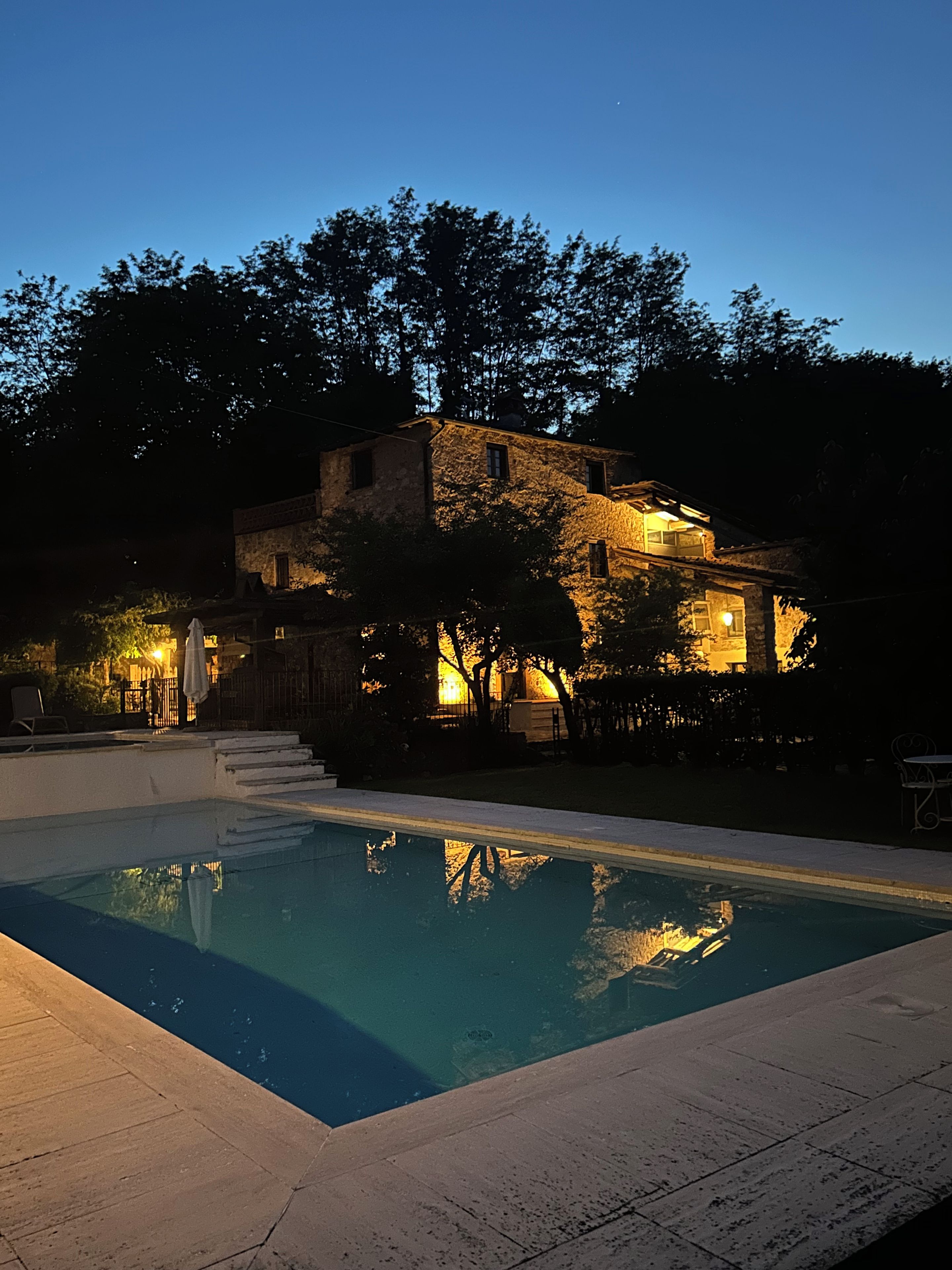 A swimming pool with a stone deck, surrounded by trees and a stone building with multiple windows and a staircase leading to the upper floor.