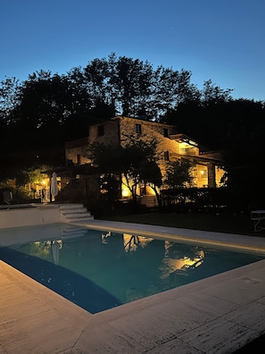 A swimming pool with a stone deck, surrounded by trees and a stone building with multiple windows and a staircase leading to the upper floor.