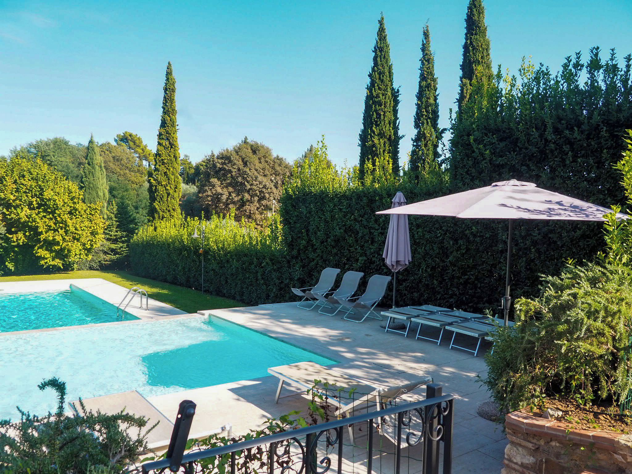 A rectangular swimming pool with a clear blue water, surrounded by a paved deck. There are lounge chairs and a covered umbrella area.