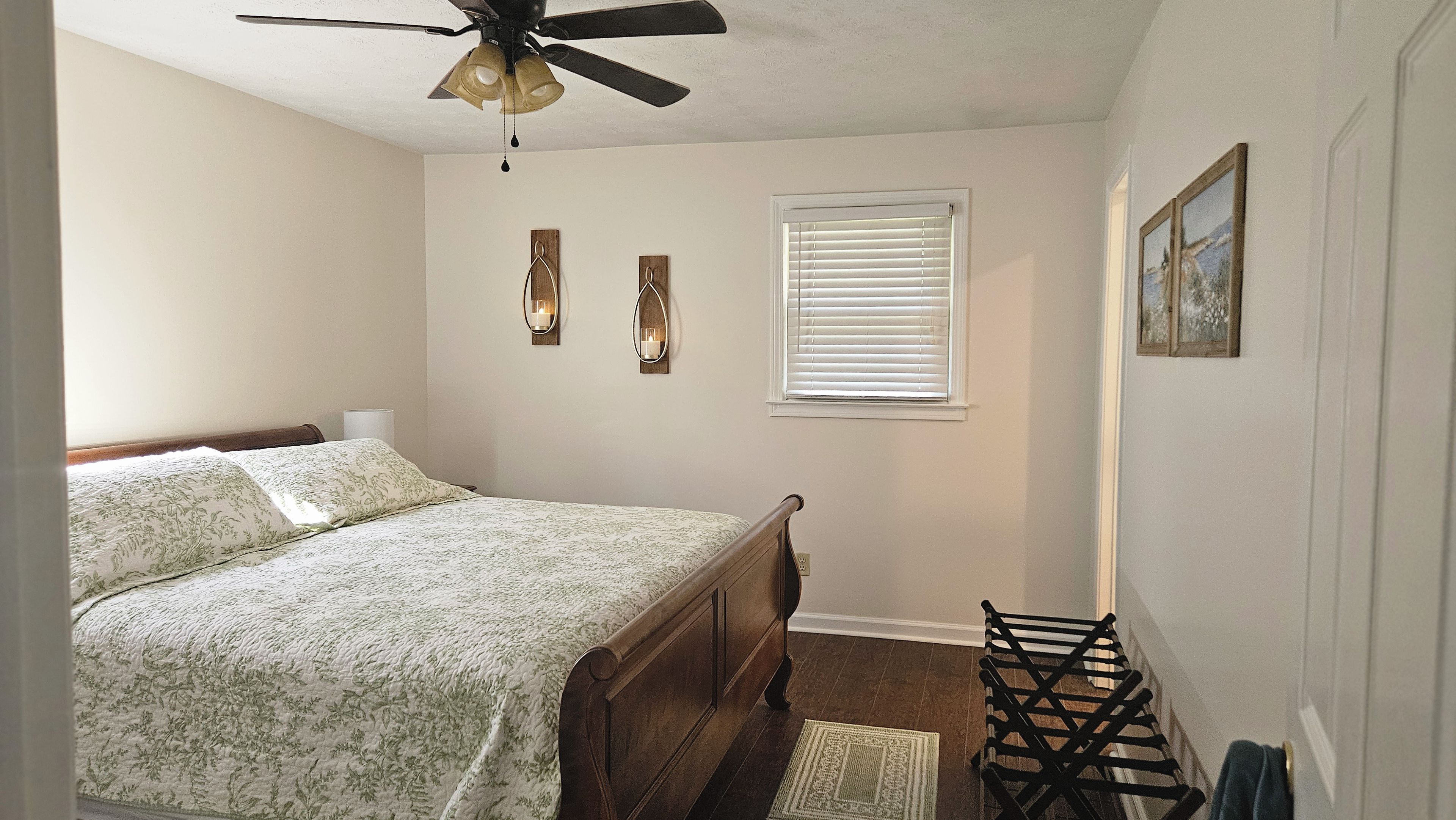 A bedroom with a bed, a ceiling fan, a window with blinds, and a wall-mounted light fixture.