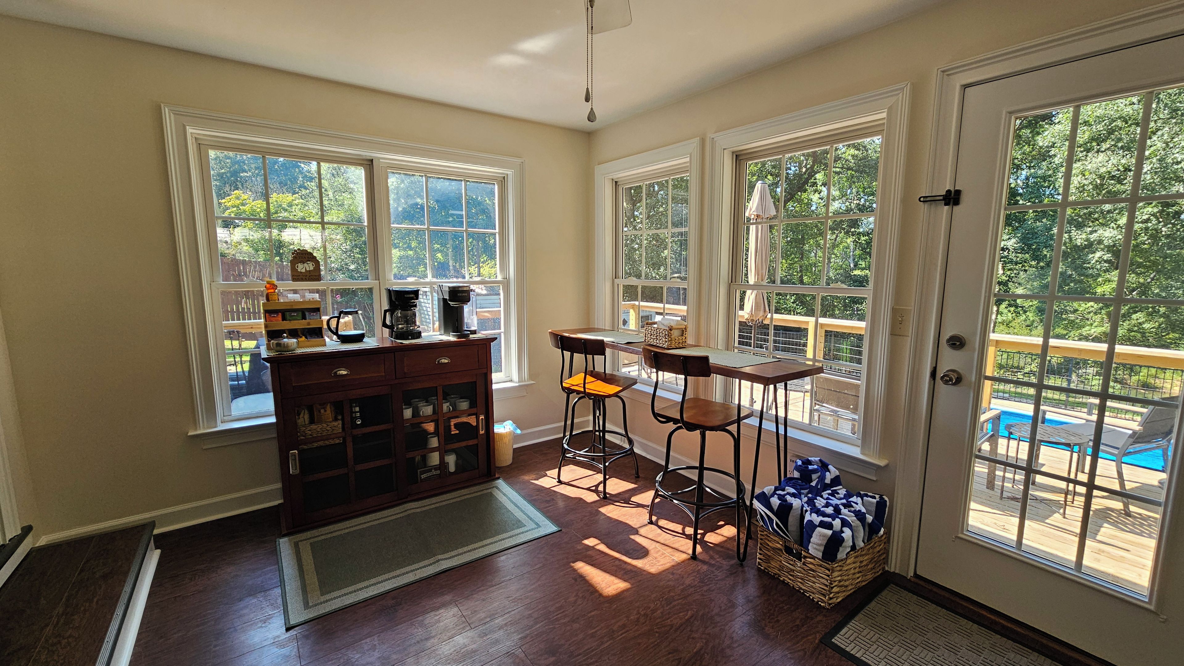 A sunlit room with a wooden cabinet, two bar stools, a small table, and a view of a swimming pool through large windows.