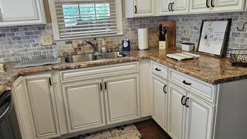 Una cocina con armarios blancos, encimera de granito, ventana con persianas y suelo de madera.