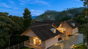 A two-story wooden cabin with a balcony, surrounded by lush greenery and a hilly landscape.