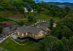 A large, two-story wooden house with a balcony, surrounded by a fenced property and lush greenery.