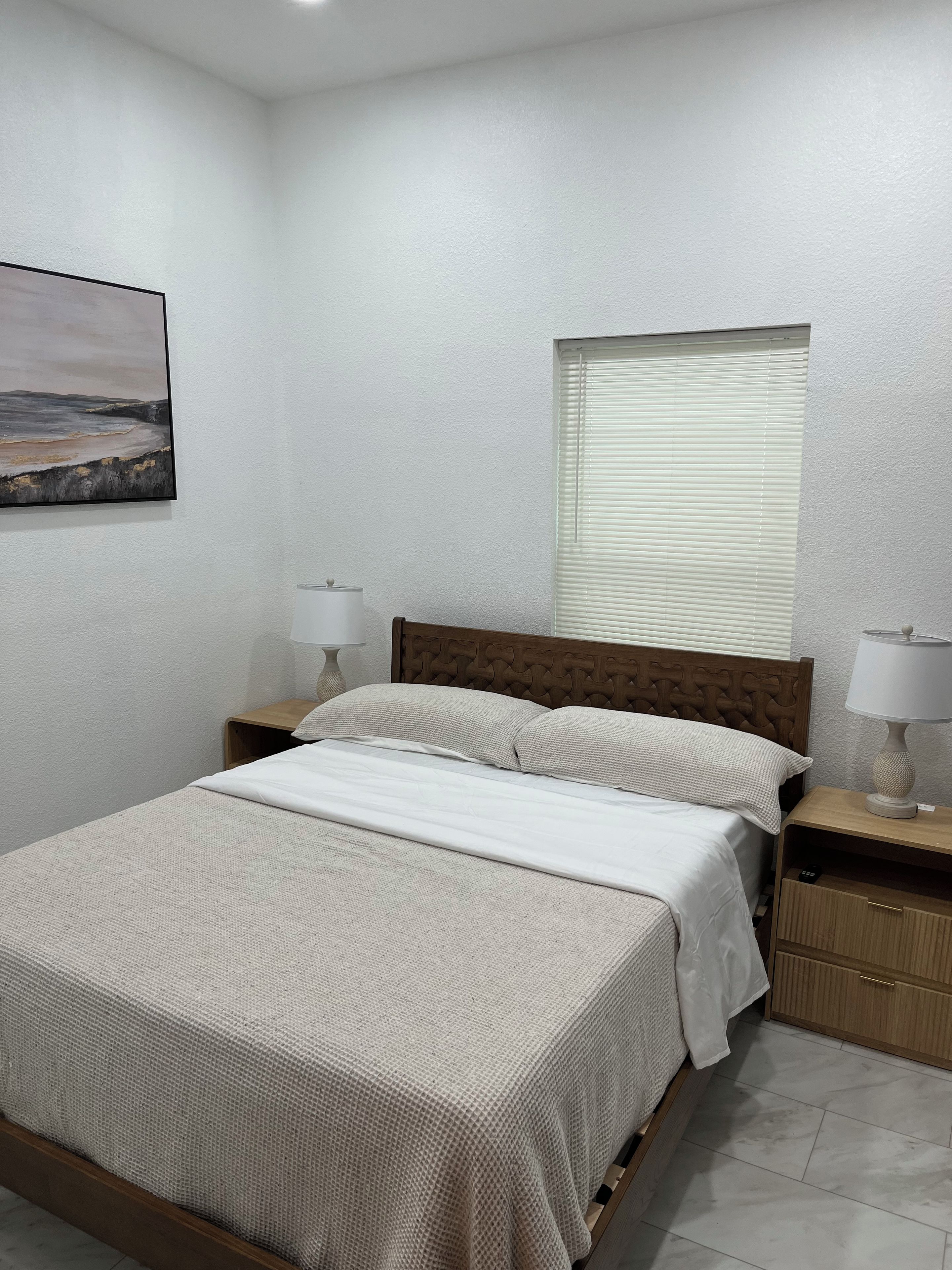 A bedroom with a bed, nightstands, a lamp, a window with blinds, and a painting on the wall.