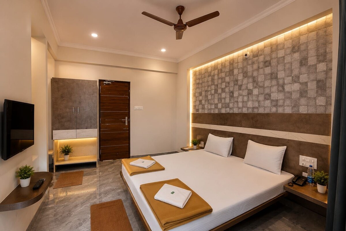 A modern hotel room with a large bed, a flat-screen TV mounted on the wall, a ceiling fan, and a decorative wall panel.