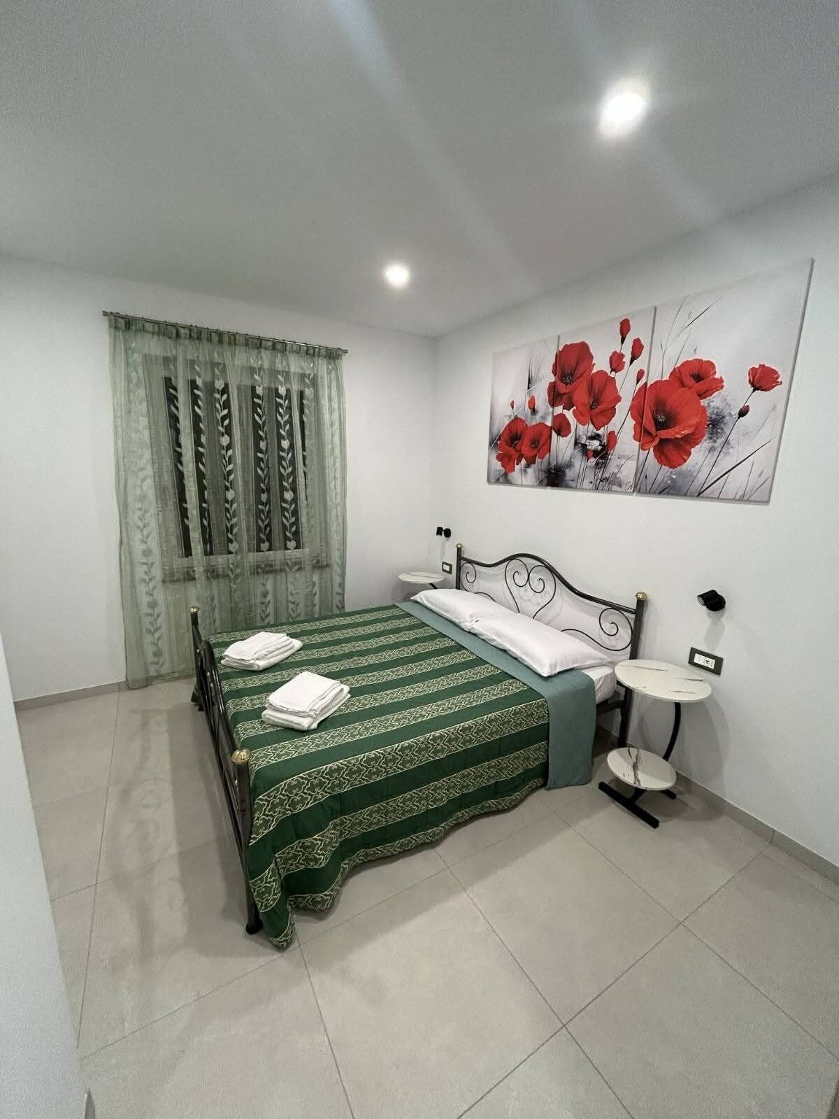 Een slaapkamer met een bed, een nachtkastje, een wandlamp en een schilderij van rode bloemen.