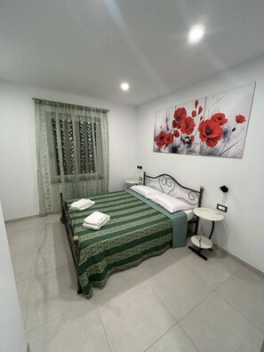 Una camera da letto con un letto, un comodino, una lampada a parete e un quadro con fiori rossi.