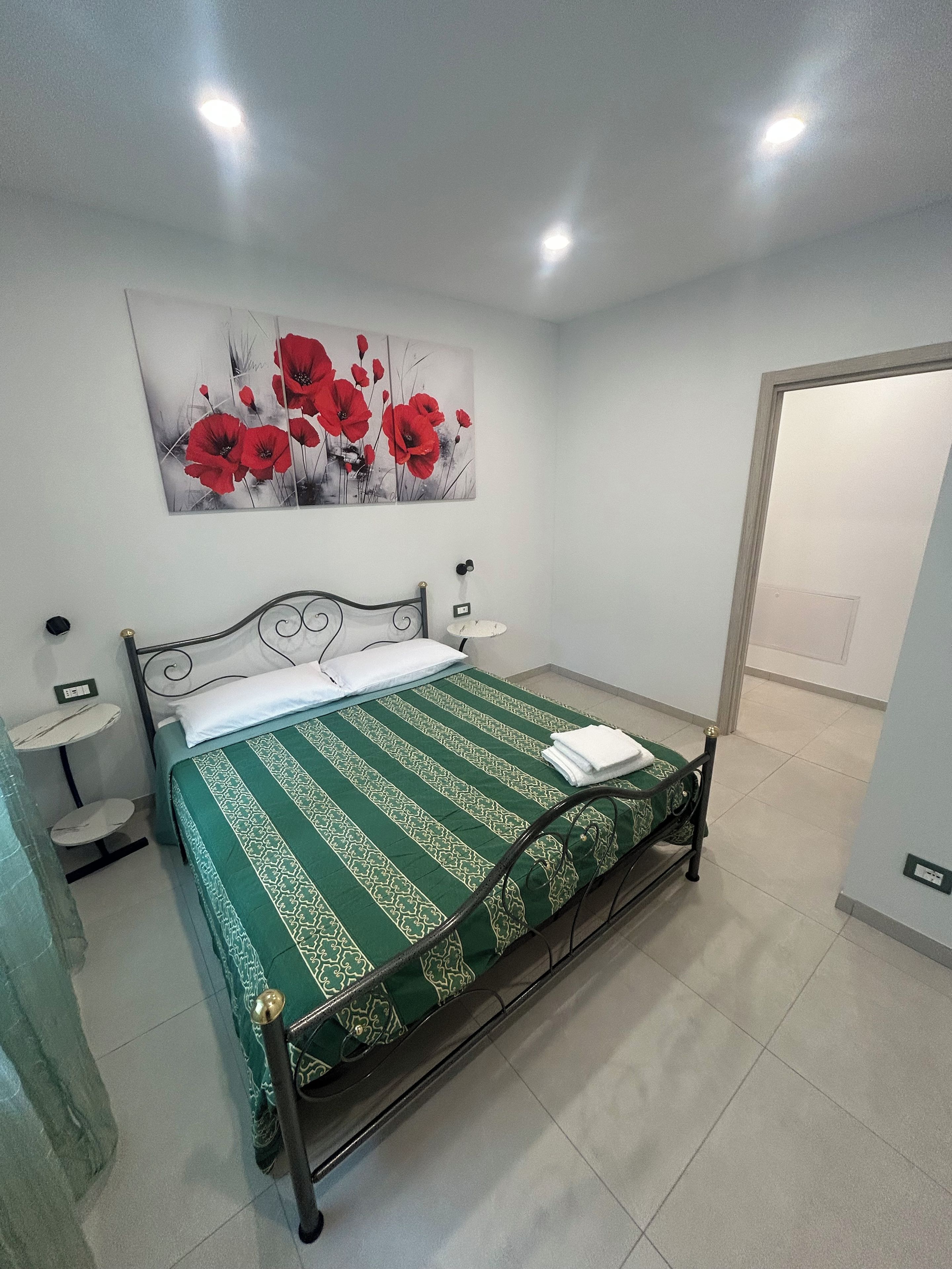 A neatly made bed with a green and white patterned bedspread, a wall-mounted painting of red flowers, and a small bedside table with a lamp.