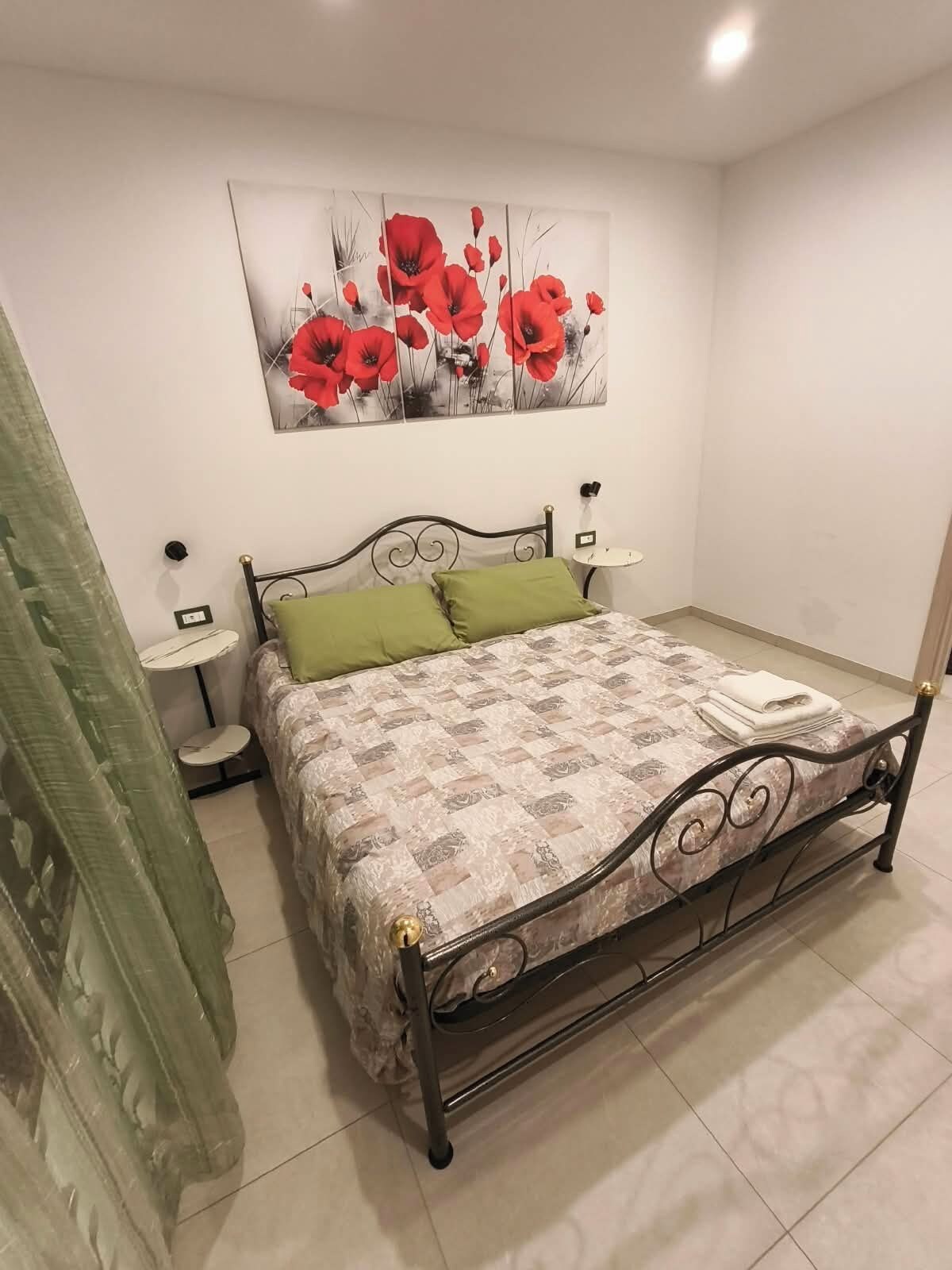 Ein Schlafzimmer mit einem Bett, grünen Kissen, einem Nachttisch, einem Wandbild und einem Vorhang.