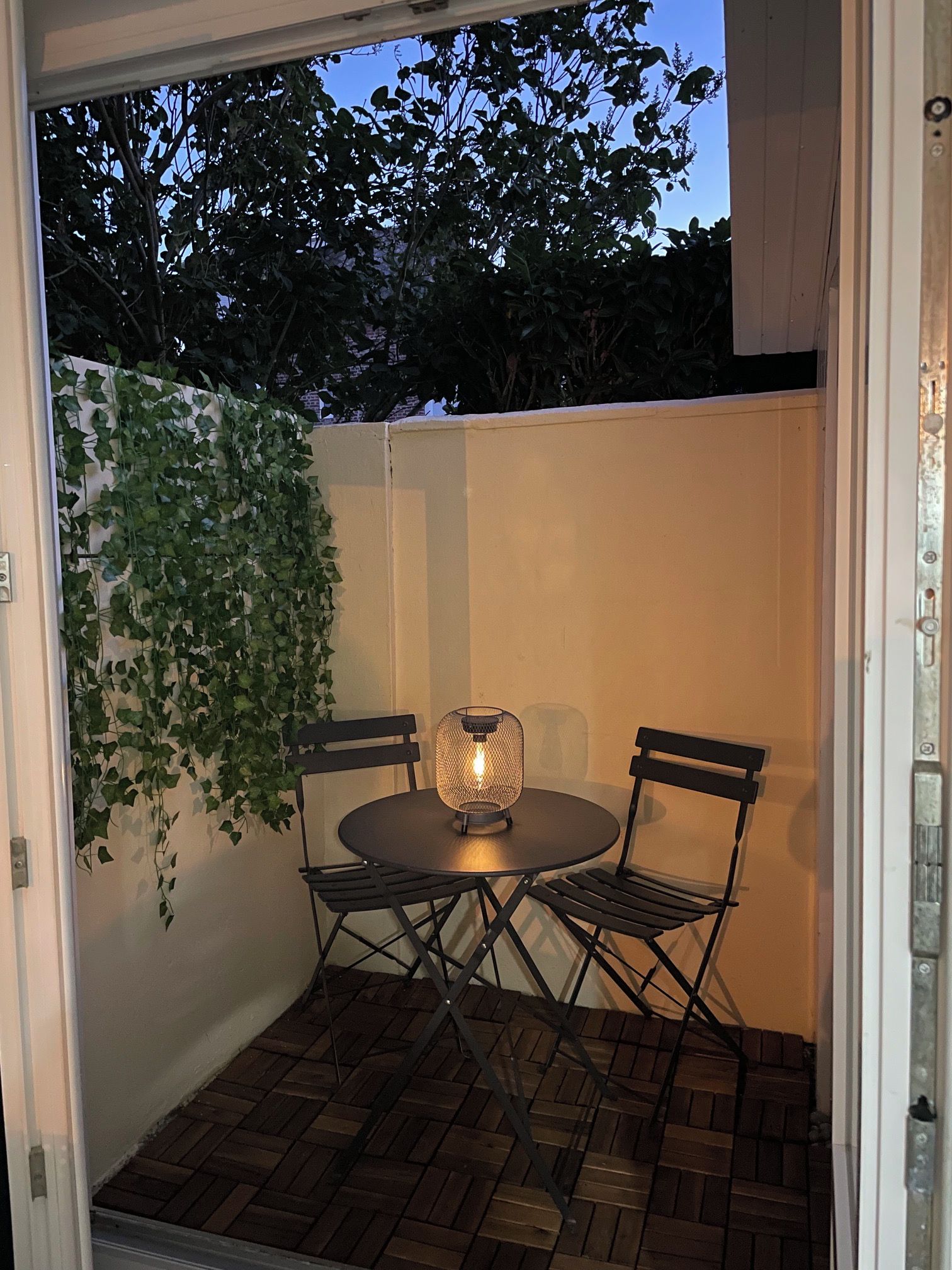 Une terrasse extérieure avec une table ronde et deux chaises, une plante suspendue et une lanterne.