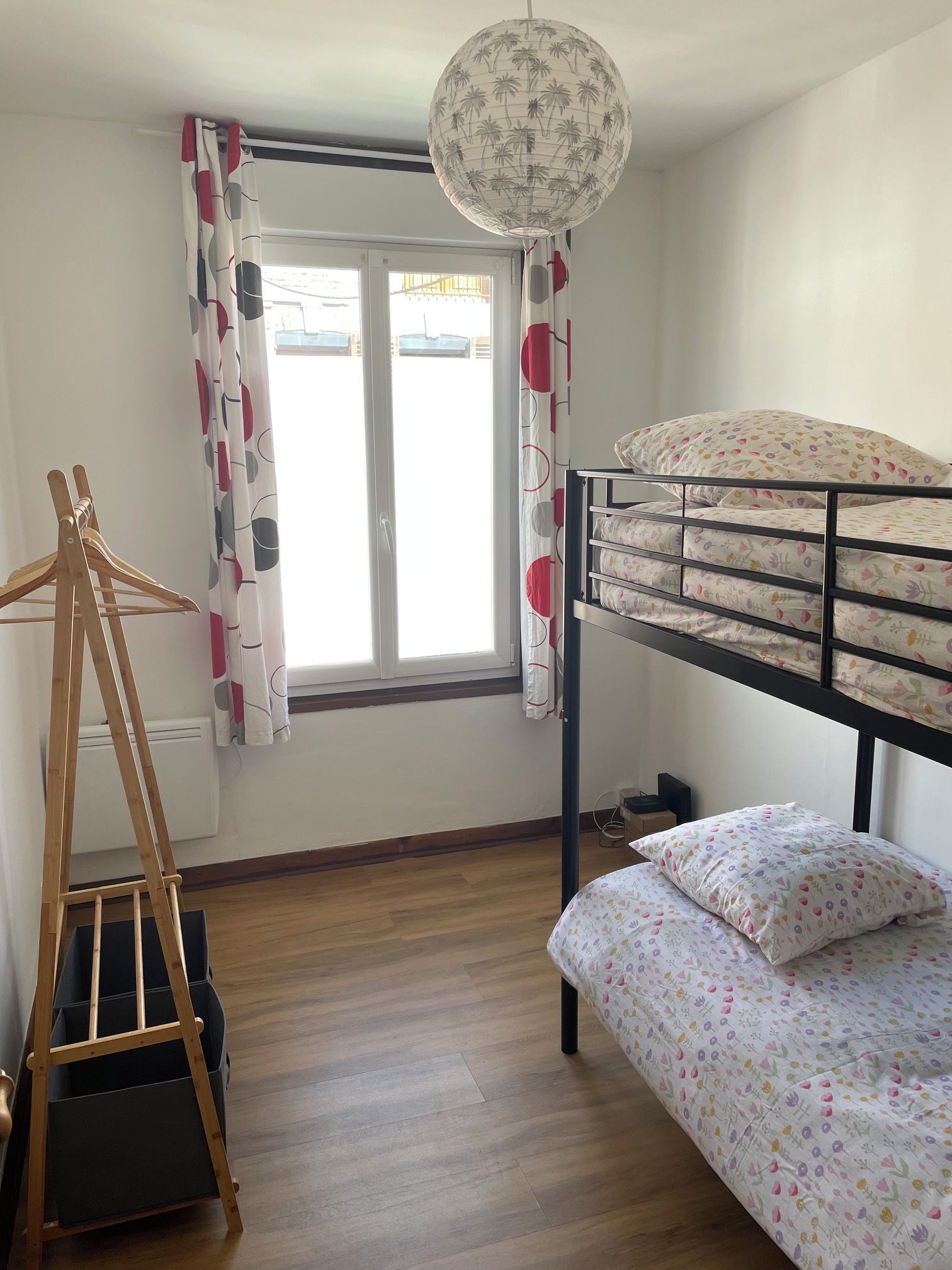 Une chambre avec un lit superposé, un porte-manteau en bois, une lampe en forme de globe et une fenêtre avec des rideaux.