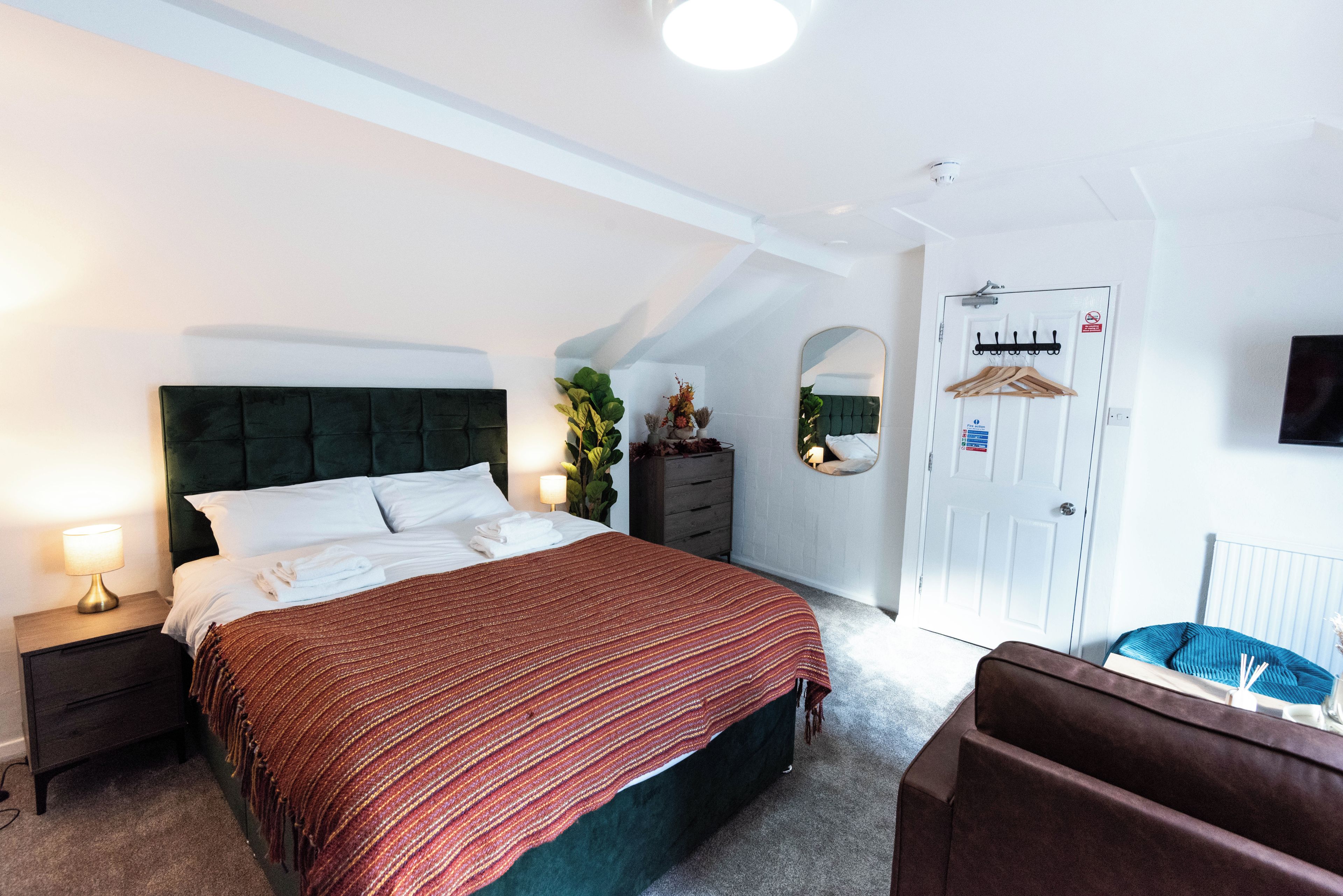 A hotel room with a bed, a nightstand, a dresser, a mirror, a coat rack, and a door with a fire safety sign.