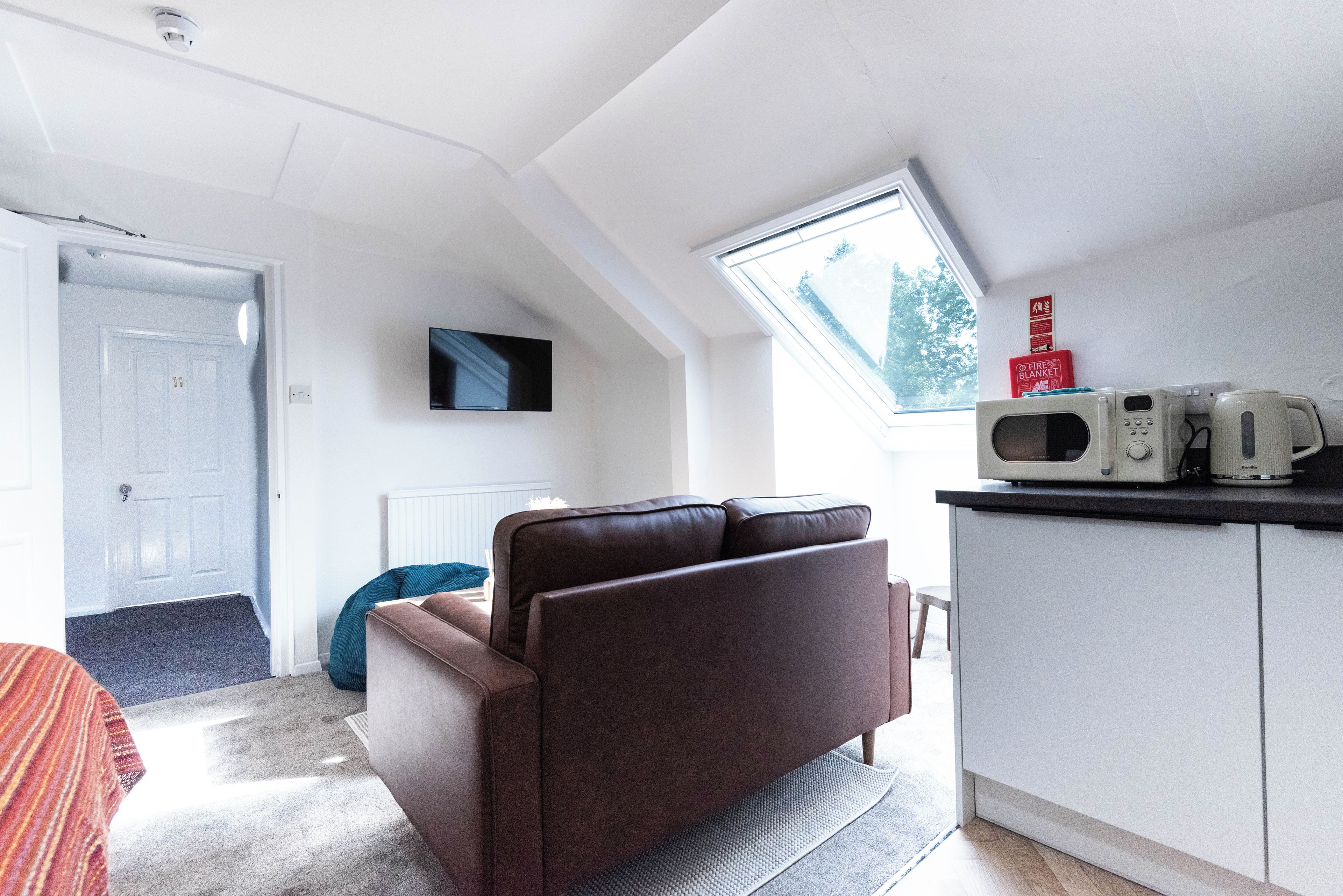 A modern living room with a brown sofa, a television, and a kitchen area with a microwave and kettle.
