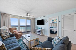A hotel room with a balcony, a flat-screen TV, a bed, and a seating area with a coffee table. - Pelican Beach 1516 2 Bedroom Condo (Destin)