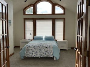 Une chambre à coucher avec un grand lit, deux tables de chevet, une fenêtre avec des stores et une porte dotée d’un panneau en verre.