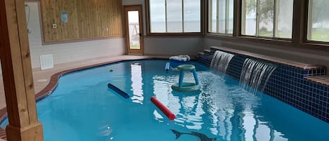 An indoor swimming pool with a wooden wall, a view of the sea, and a blue tiled edge.