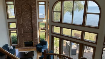 A spacious living room with a brick fireplace, multiple windows offering views of the sea, and a staircase leading to an outdoor area.