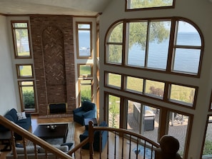 A spacious living room with a brick fireplace, multiple windows offering views of the sea, and a staircase leading to an outdoor area.