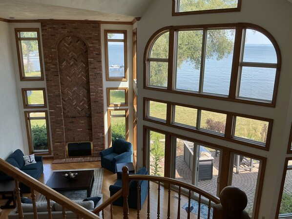 A spacious living room with a brick fireplace, multiple windows offering views of the sea, and a staircase leading to an outdoor area.