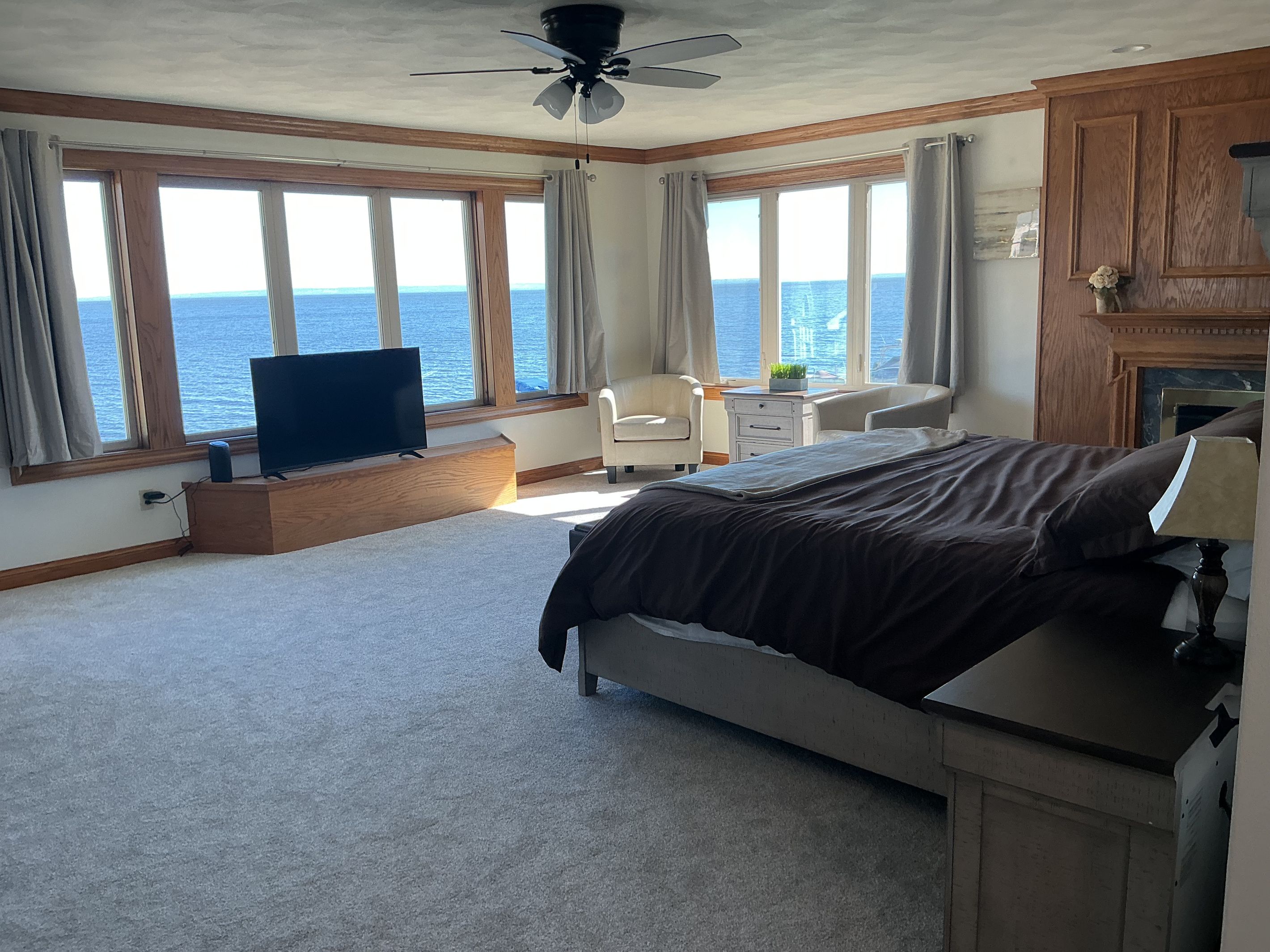 A bedroom with a bed, a desk, a chair, a television, and large windows with a view of the sea.