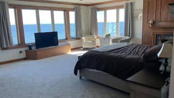 A bedroom with a bed, a desk, a chair, a television, and large windows with a view of the sea.